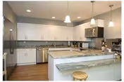 Modern kitchen with white cabinets, granite countertops, stainless steel appliances including a refrigerator, dishwasher, and microwave. There are three pendant lights hanging above a kitchen island with a wooden stool. The floor is wooden and the backsplash is a light-colored tile.
