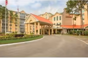 Front exterior view of a multi-story senior living facility named Plaza West with a covered entrance, landscaped greenery, and an American flag on a flagpole.