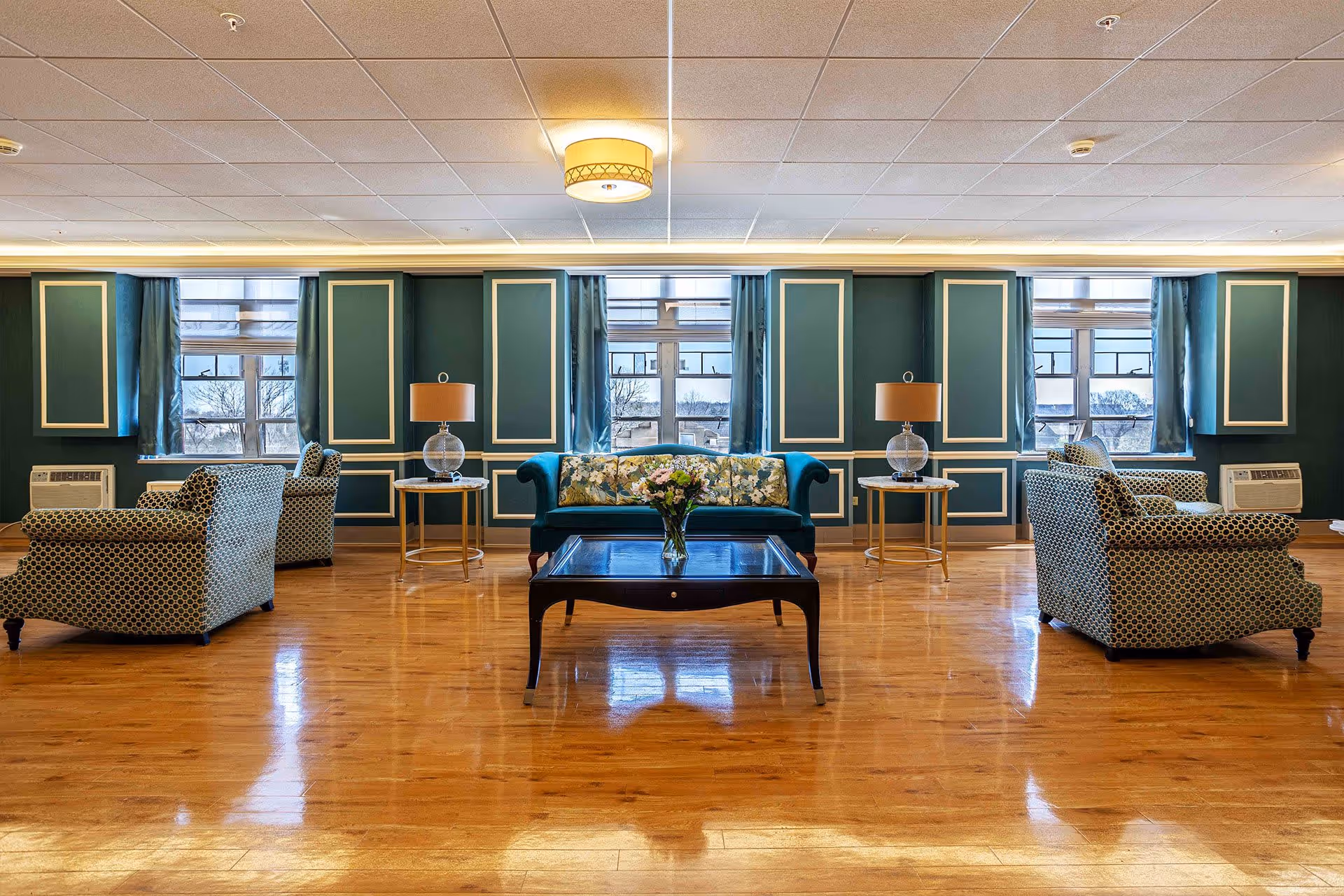 A spacious and elegant living room area with polished wooden floors, teal walls with white trim, and multiple windows letting in natural light. The room features a teal upholstered sofa with floral cushions, two patterned armchairs, two side tables with glass lamps, and a dark wooden coffee table with a vase of flowers in the center.