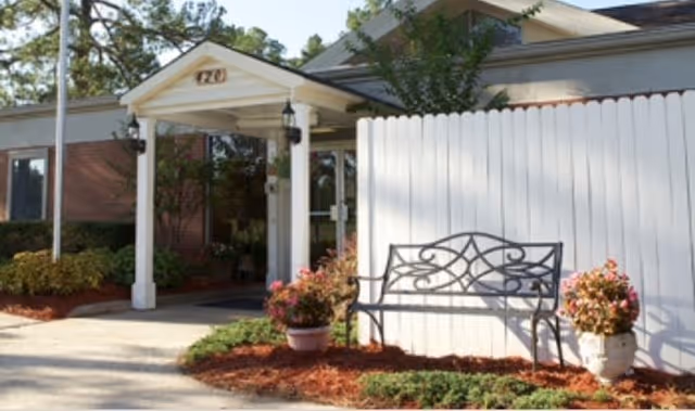 Entrance to a building with a white covered porch supported by columns, a white picket fence, a decorative black metal bench, and potted plants with flowers on either side of the bench. The building is surrounded by greenery and trees.