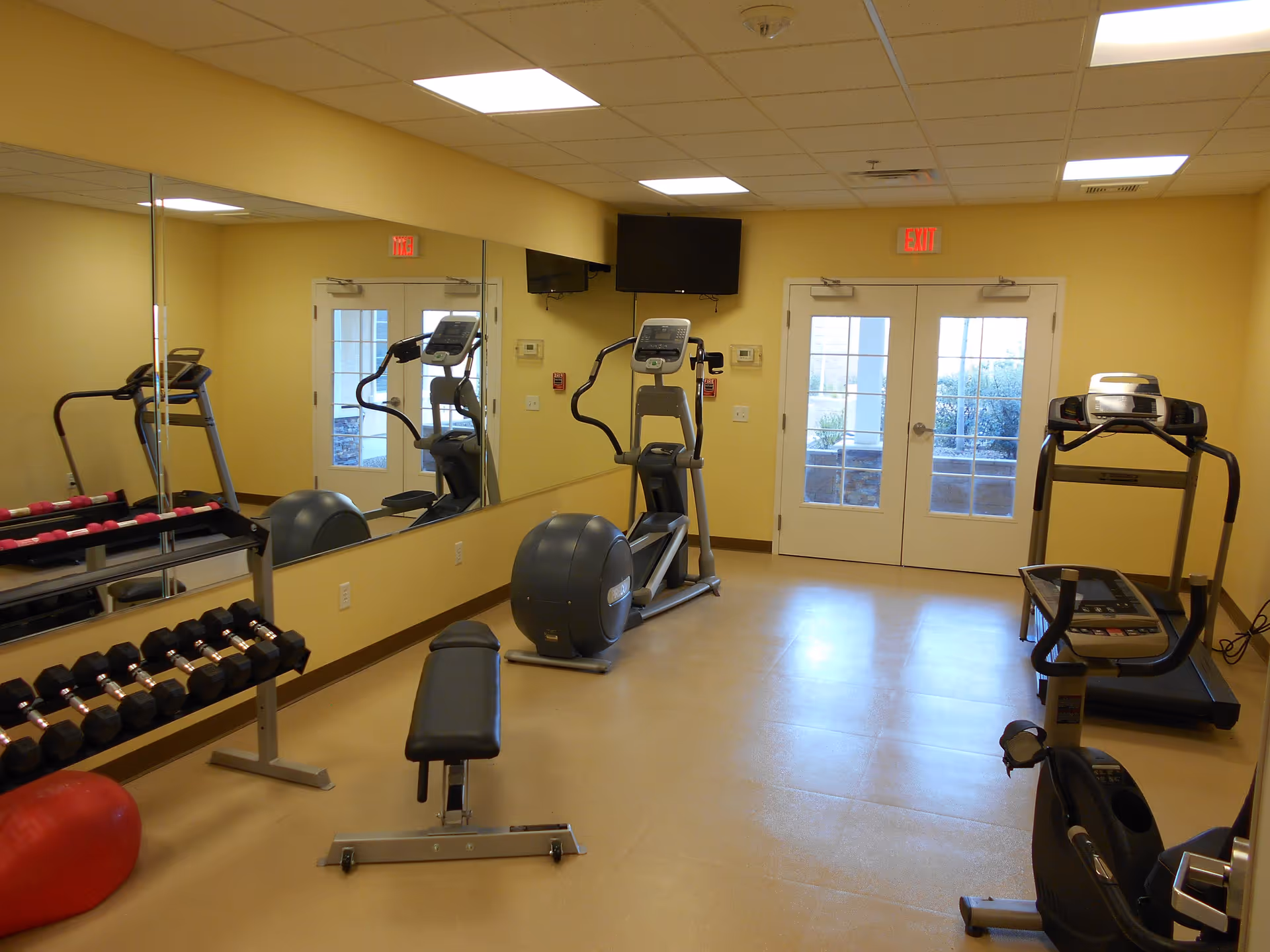 A small fitness room with exercise equipment including a treadmill, elliptical machine, stationary bike, weight bench, and dumbbells. The room has yellow walls, a large mirror on one side, and double glass doors leading outside. There is a wall-mounted TV above the elliptical machine.