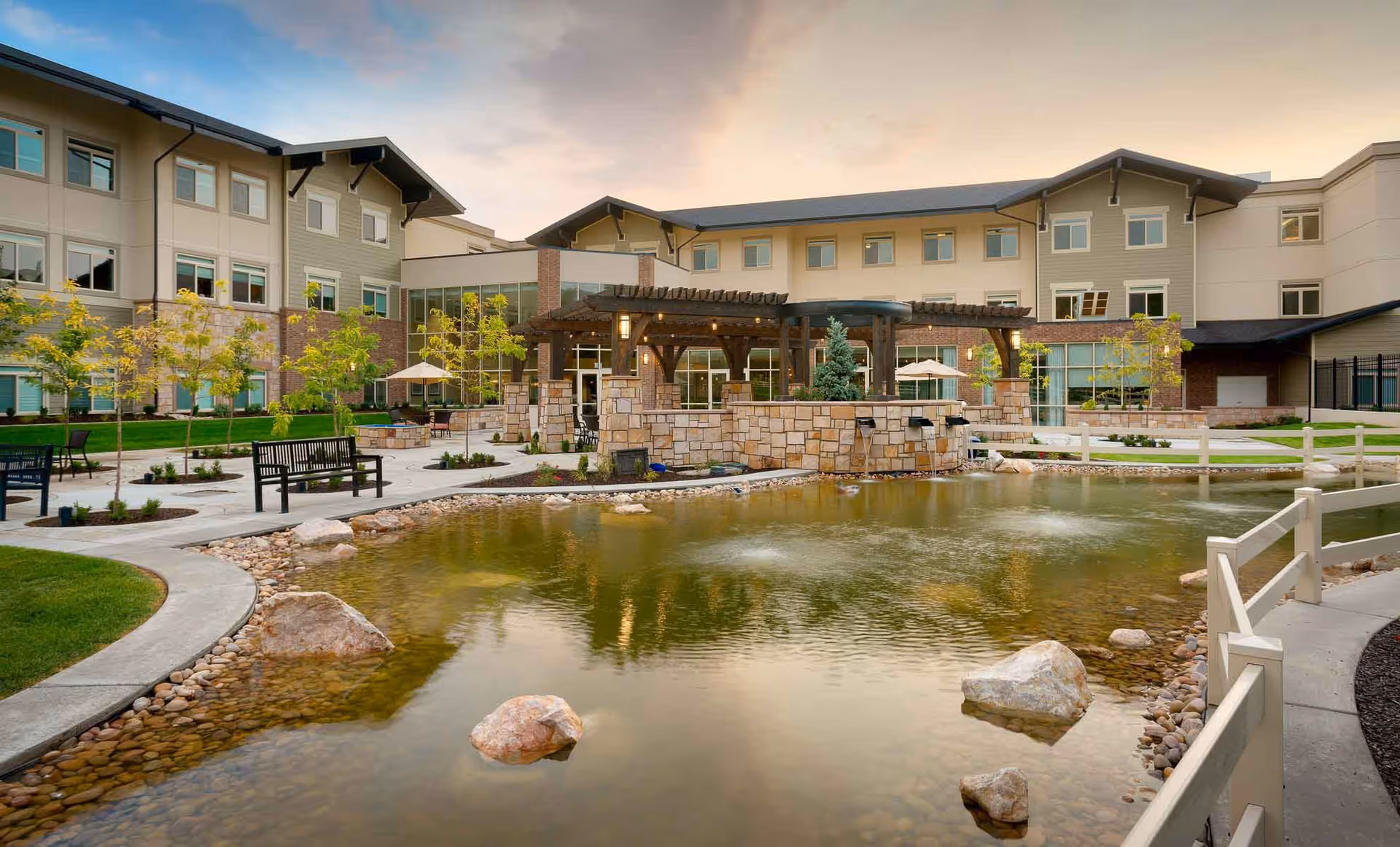 Outdoor courtyard featuring a pond, benches, pergola, and landscaping in front of a multi-story senior living building.