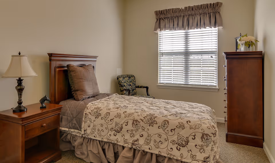 A cozy single bedroom with a wooden bed, patterned comforter, nightstand with lamp, armchair, window with blinds, and a tall dresser.