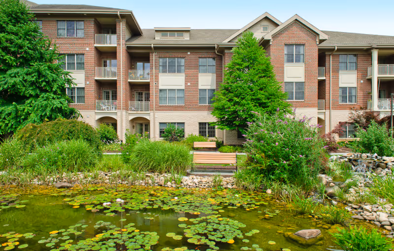 Three-story brick residential building with balconies overlooking a landscaped pond with lily pads, benches, and greenery.