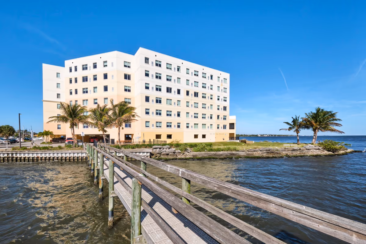 A multi-story senior living building sits along a waterfront with a wooden pier and palm trees.