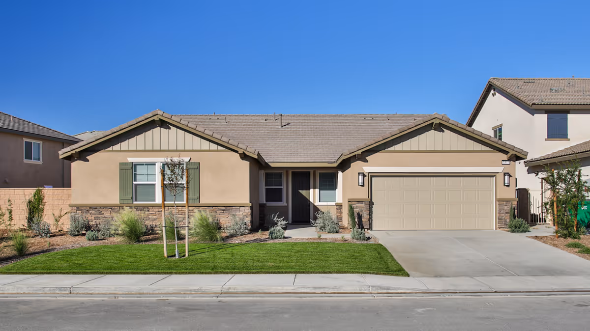Single-story beige house with an attached two-car garage, small landscaped front yard, and clear blue sky.