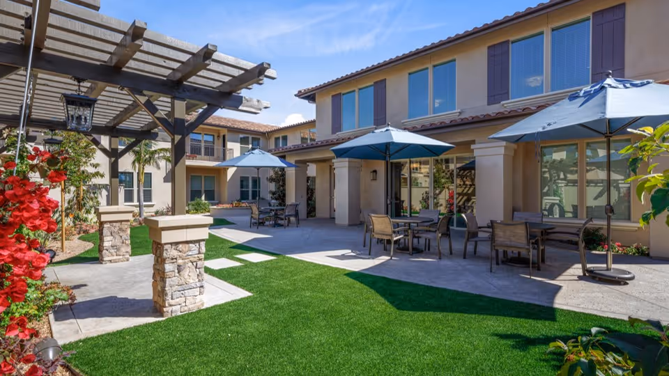 Outdoor courtyard area of a senior living facility with green artificial grass, several tables with chairs and blue umbrellas, a pergola with stone pillars, and a two-story beige building with multiple windows in the background under a clear blue sky.