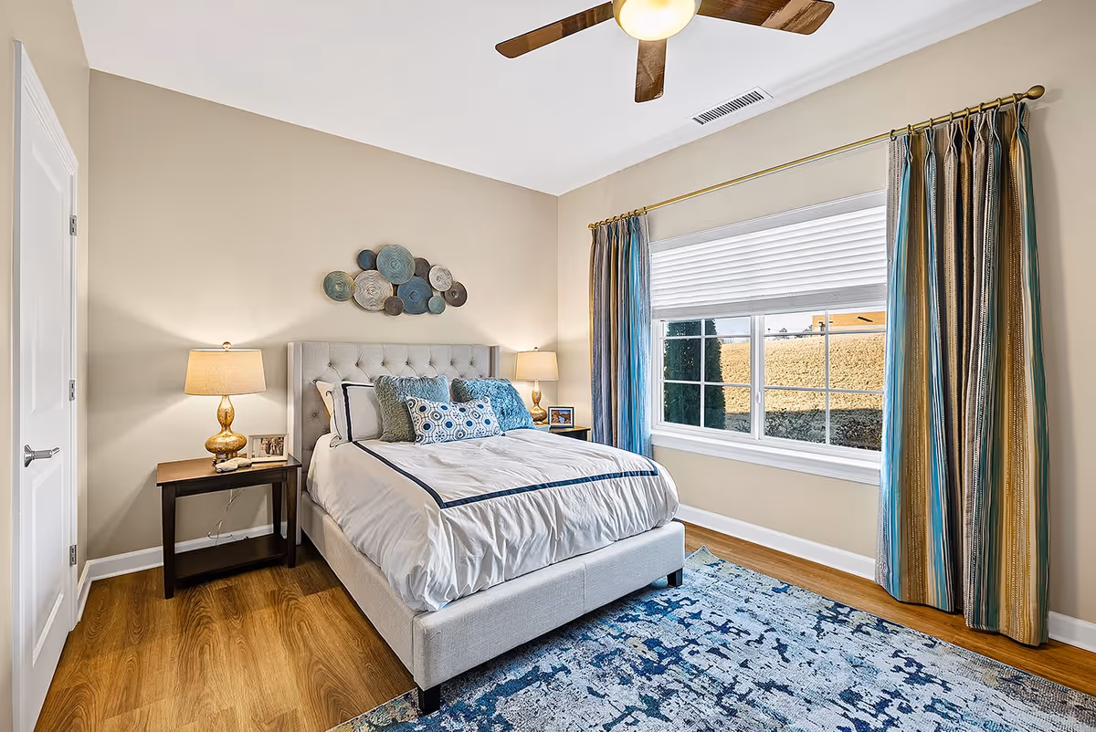 A bright bedroom with a large window covered by blue and gold striped curtains. The room features a bed with a tufted headboard, white bedding with blue accents, and decorative pillows. There are two bedside tables with matching lamps and framed photos. A ceiling fan with wooden blades is mounted on the white ceiling, and a blue patterned rug covers part of the wooden floor.