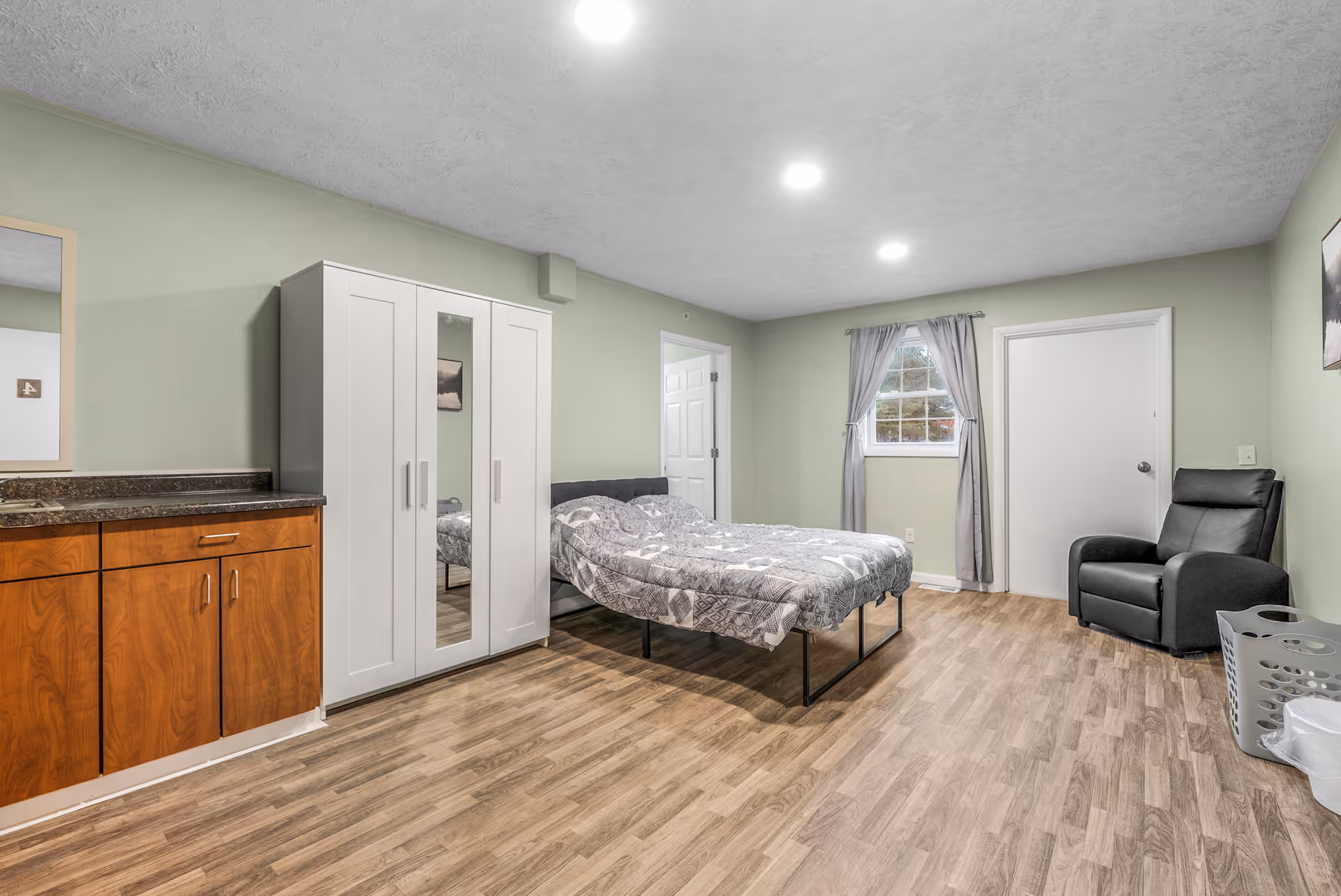 Spacious furnished bedroom with a bed, white wardrobe, kitchenette cabinets, a recliner chair, and a window.
