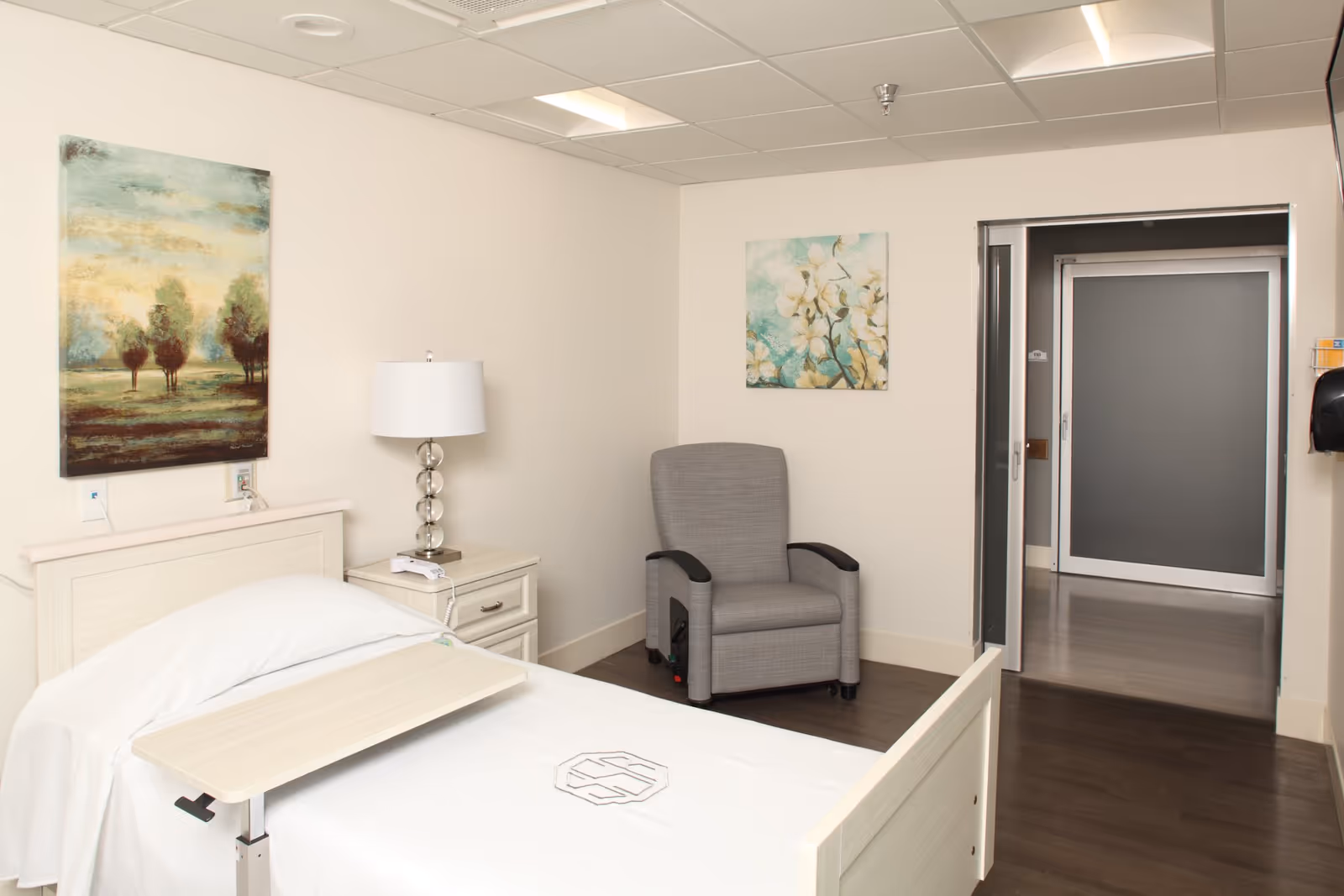 A clean and simple senior living bedroom with a single bed featuring a white bedspread and an adjustable overbed table. Next to the bed is a nightstand with a modern lamp and a telephone. A gray upholstered armchair is placed in the corner of the room. The walls are decorated with two paintings, one depicting trees and the other white flowers. The room has a doorway leading to a hallway with a closed door visible.