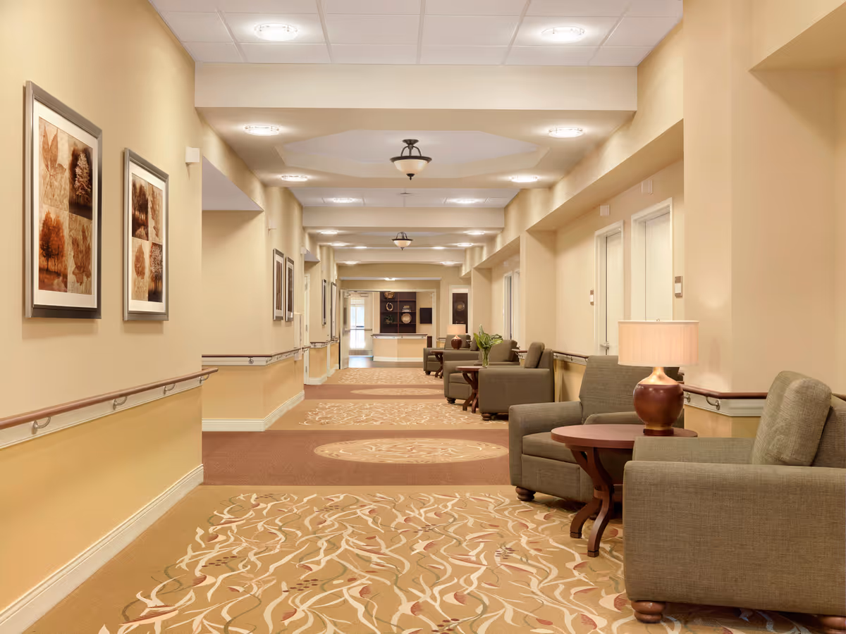A well-lit hallway in a senior living facility with beige walls and patterned carpet. The hallway features several comfortable armchairs and small tables with lamps along the right side. Handrails run along both sides of the hallway, and framed artwork is hung on the left wall. Ceiling lights are evenly spaced along the corridor, leading to a distant area with more seating and decor.