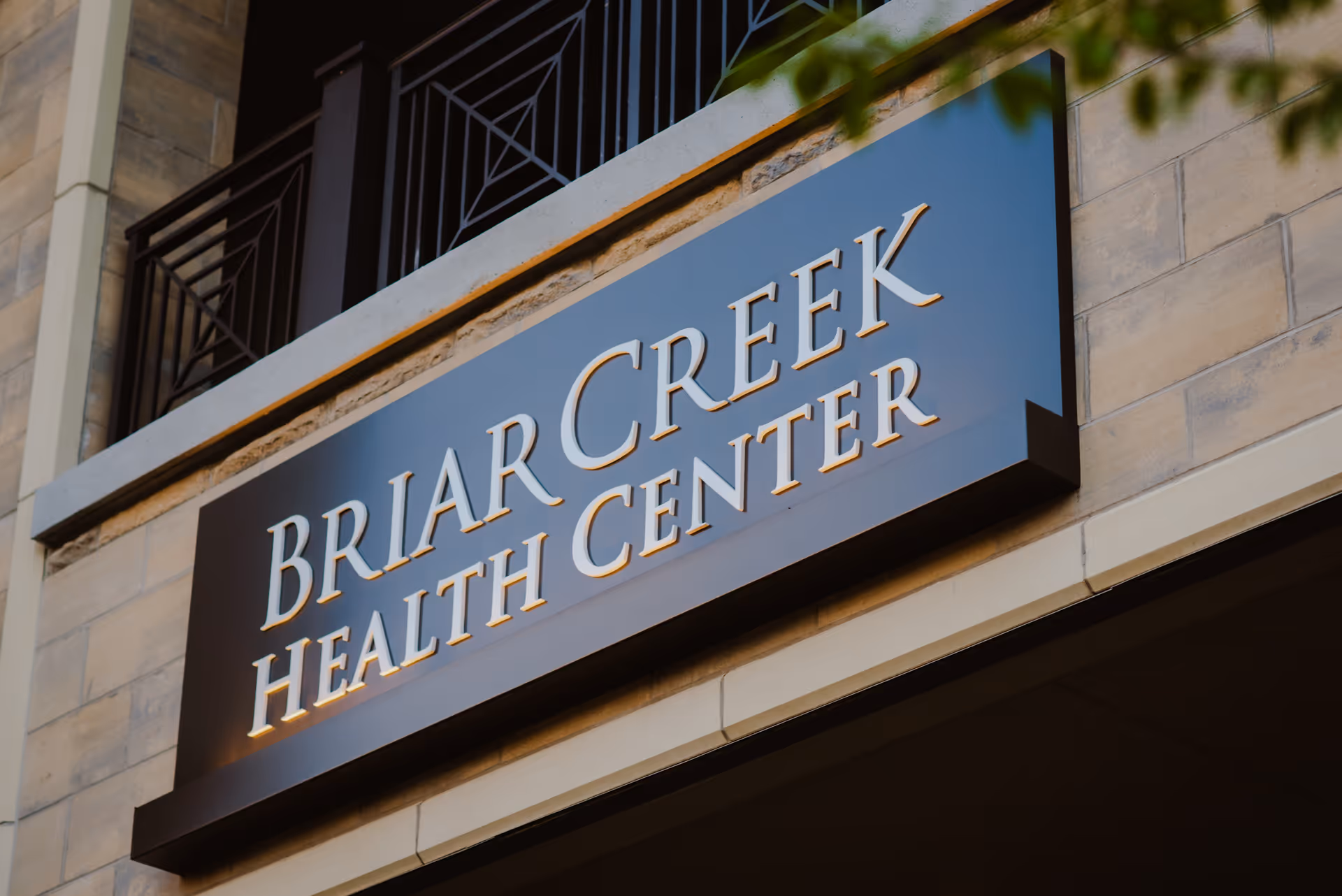 Close-up view of a building sign that reads 'Briar Creek Health Center' mounted on a stone wall with a metal railing above it.