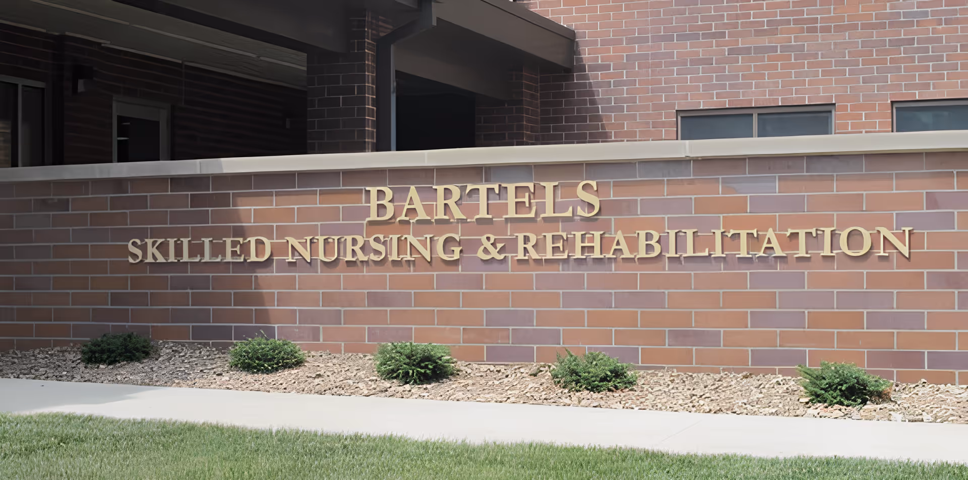 Brick exterior wall with gold letters reading 'BARTELS SKILLED NURSING & REHABILITATION', small shrubs and a sidewalk in front.