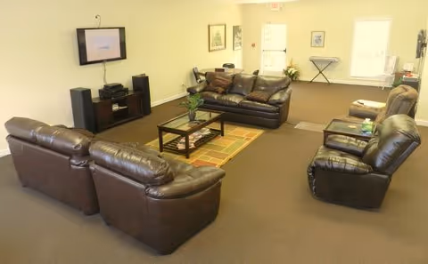 A spacious living room area with multiple brown leather sofas and armchairs arranged around a coffee table on a patterned rug. A flat-screen TV is mounted on the wall with speakers and media equipment below it. The room has beige walls, carpeted flooring, framed pictures on the walls, and windows letting in natural light.