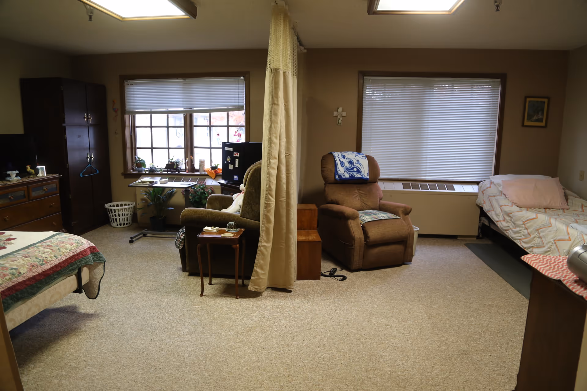 Interior view of a senior living facility room at Cumbernauld Village featuring two beds separated by a privacy curtain, a brown recliner chair with a blue and white blanket draped over it, a wooden dresser, a small table, a window with blinds, and various plants and decorations.