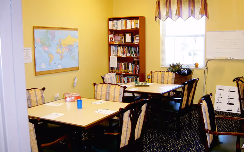 A small meeting or activity room with two square tables pushed together surrounded by eight upholstered chairs. On the tables are papers and a Yahtzee game box. A bookshelf filled with books is against the wall, and a world map is mounted on another wall. A window with patterned valances lets in natural light, and a whiteboard is visible near the window.