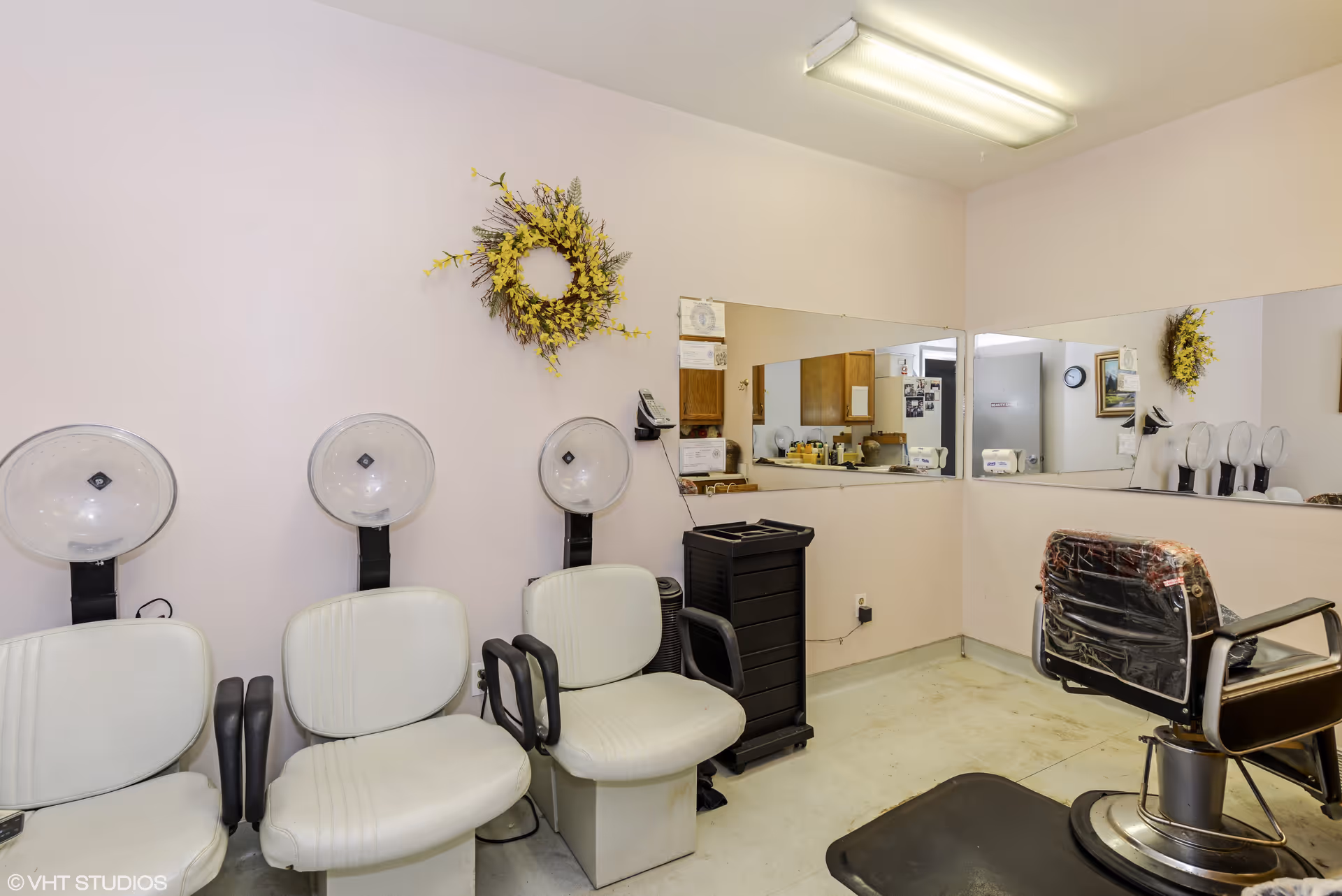 Interior of a salon area with three white hair drying chairs lined up against a light pink wall, a black salon chair with a plastic cover, a large mirror on the wall reflecting the room, and a yellow floral wreath hanging on the wall.