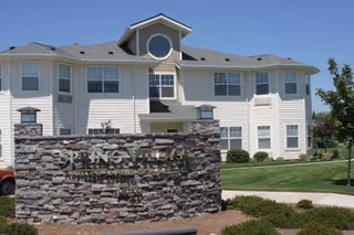 Exterior view of a two-story residential building with white siding and multiple windows under a clear blue sky. In front of the building is a large stone sign with the text 'SPRING VILLAGE' and additional smaller text below it. The area around the building is landscaped with green grass, bushes, and a tree.