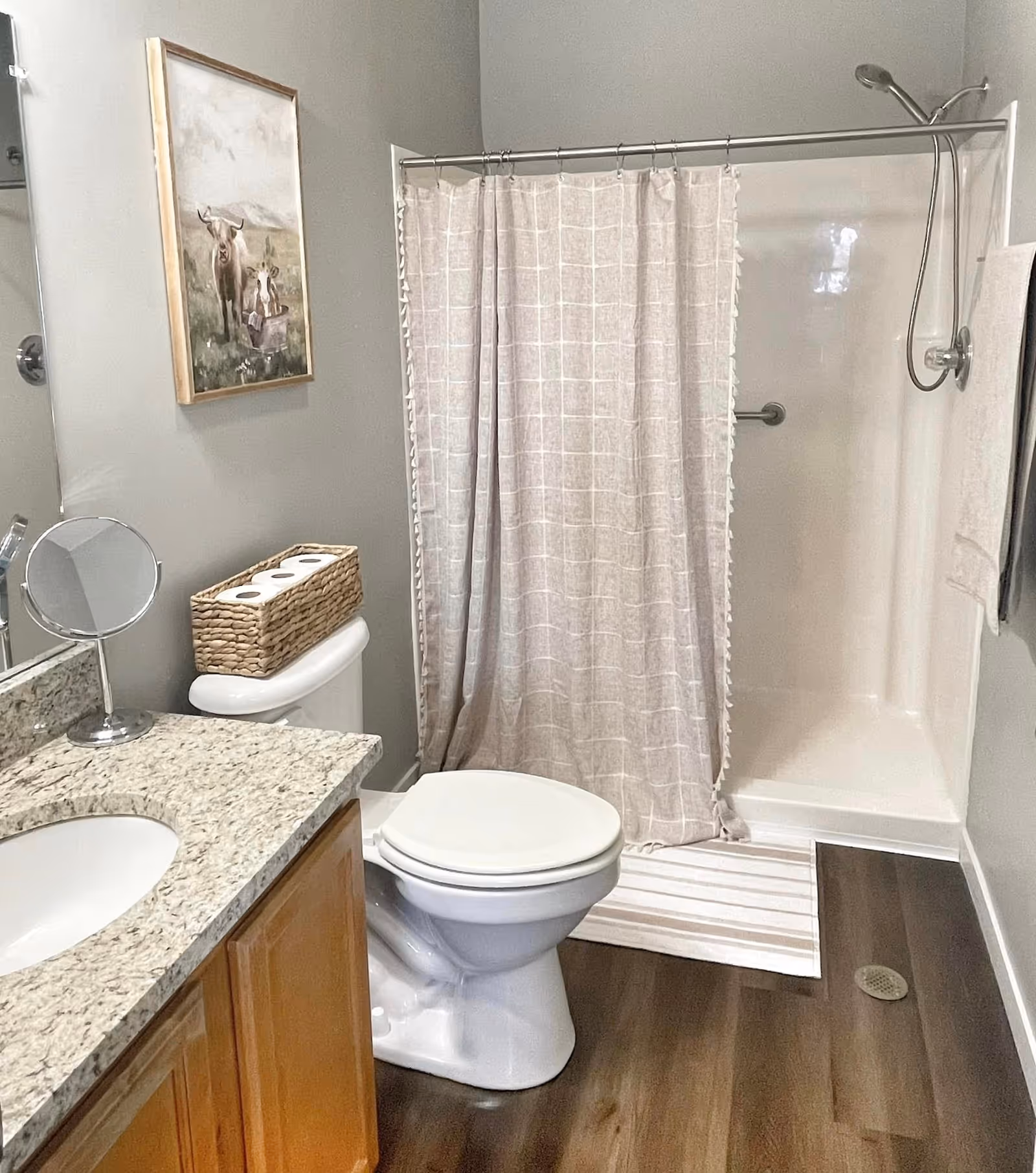 A bathroom with a granite countertop sink, wooden cabinet, white toilet with a wicker basket holding three rolls of toilet paper on top, a shower with a beige checkered curtain, a striped bath mat, and a towel hanging on the wall. There is a framed painting of two cows on the wall above the toilet.