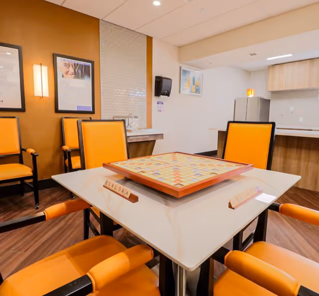 Communal game room with orange chairs around a table featuring a Scrabble board and a kitchenette in the background.