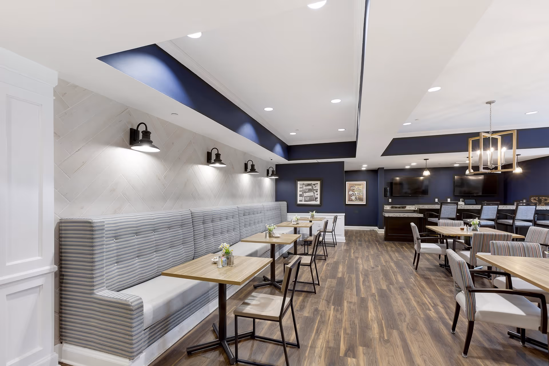 A modern dining area in a senior living facility featuring a long upholstered bench with striped fabric along one wall, several wooden tables with chairs, wood flooring, and navy blue and white walls. The ceiling has recessed lighting and decorative light fixtures, with framed artwork on the walls and a counter area with bar stools and two mounted flat-screen TVs in the background.