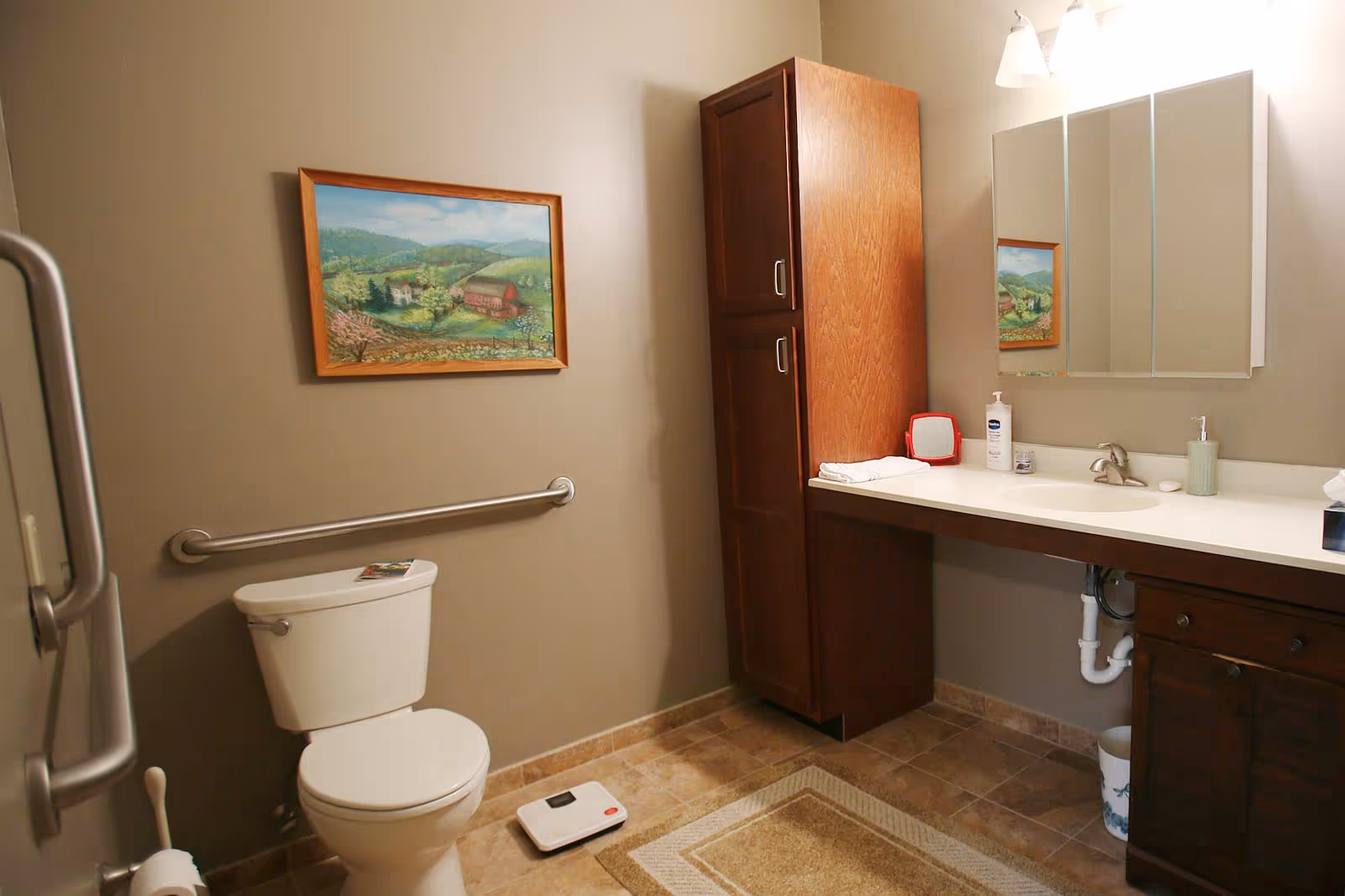 A bathroom with beige walls and tiled floor, featuring a white toilet with a grab bar on the wall beside it, a white scale on the floor, a wooden cabinet, a white countertop with a sink, a mirror above the sink, and a framed landscape painting on the wall.