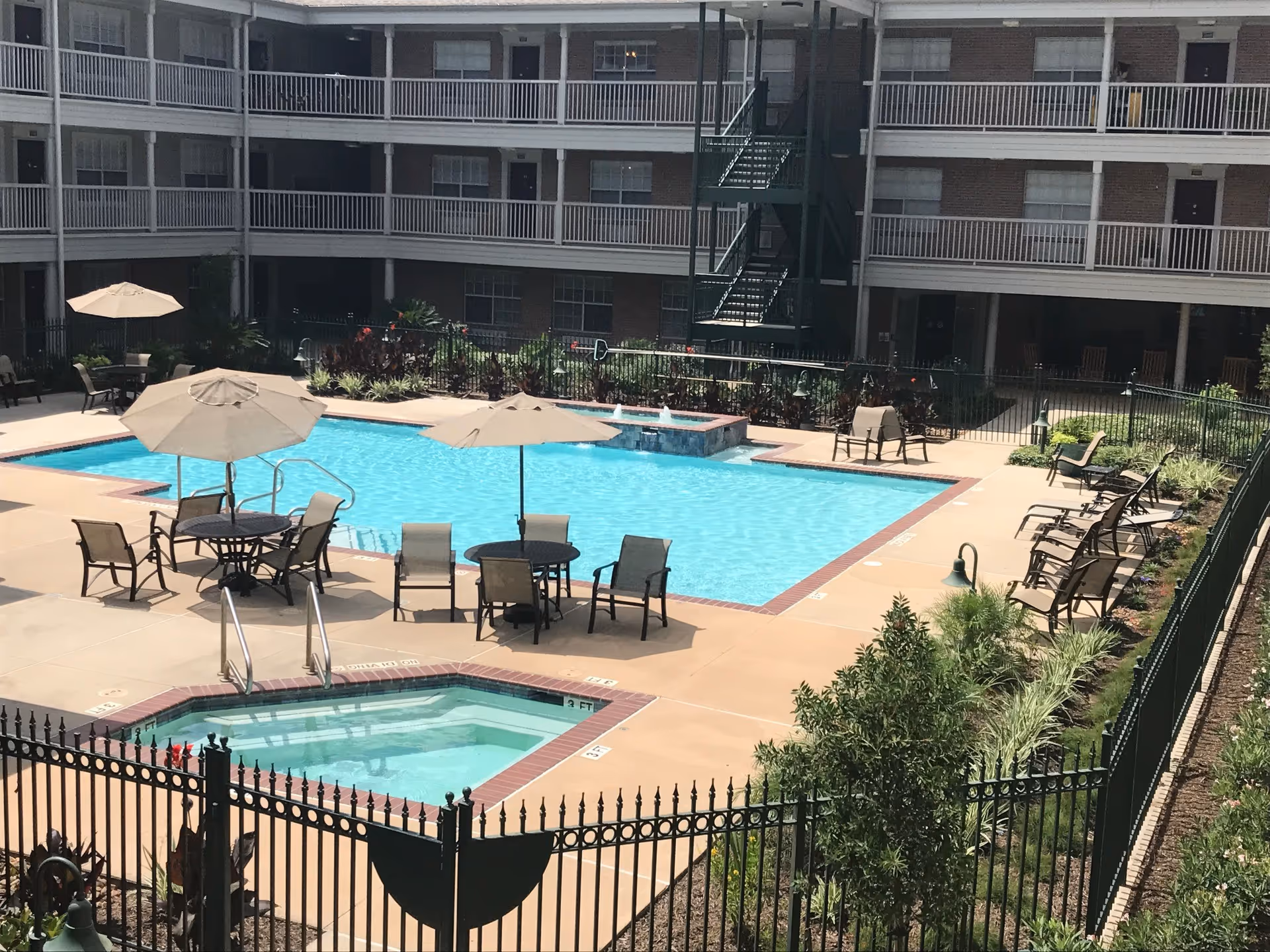 Outdoor swimming pool area at a senior living facility with several lounge chairs, tables with umbrellas, and a hot tub. The pool is surrounded by a black metal fence and landscaping, with a multi-story building in the background featuring balconies and a green fire escape.