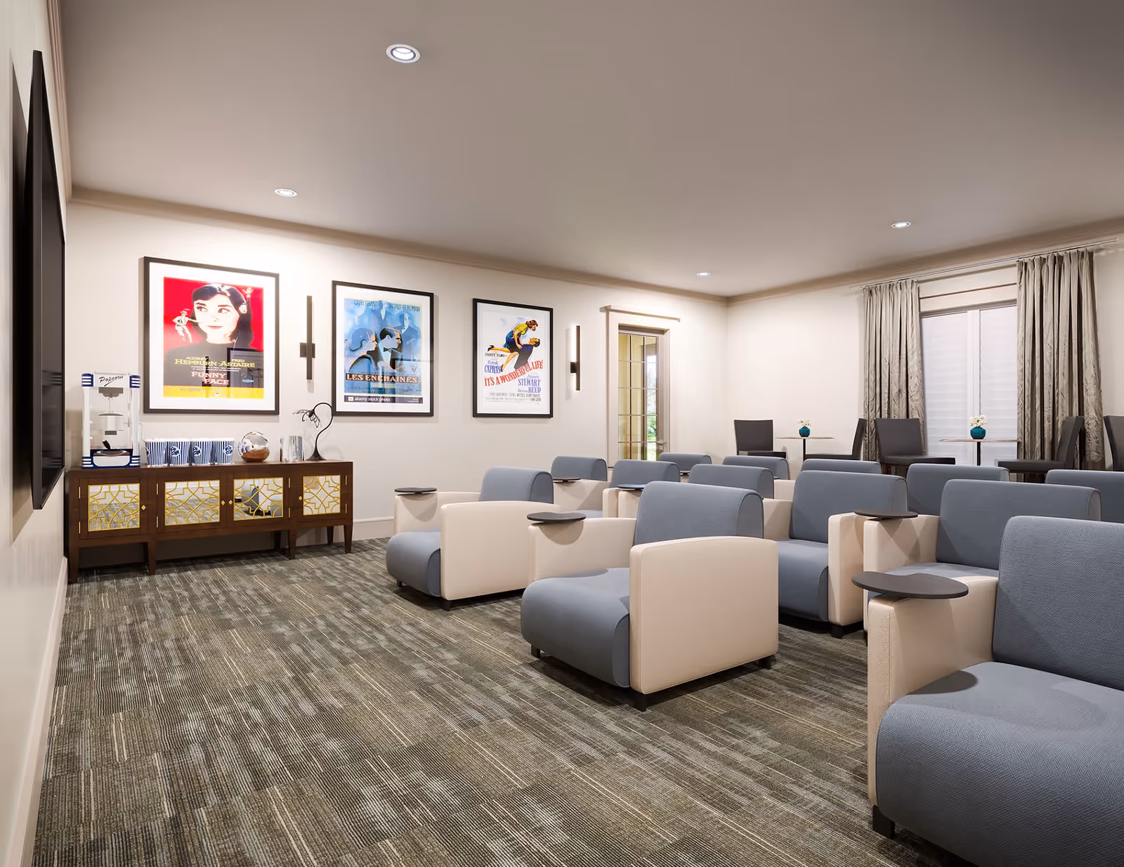 A cozy movie or presentation room with multiple rows of comfortable blue and beige armchairs, each with a small attached table. The room has a carpeted floor, three framed vintage movie posters on the wall, a wooden sideboard with a popcorn machine and cups, and large windows with curtains letting in natural light.