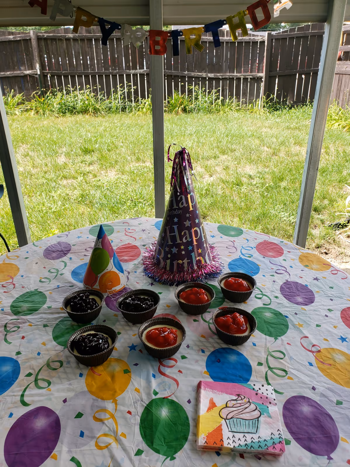 A round table covered with a colorful balloon-themed tablecloth is set up outdoors with small cups of chocolate and strawberry pudding, two birthday party hats, and a stack of napkins with a cupcake design. A 'Happy Birthday' banner is hanging on the screened porch behind the table, with a grassy backyard and wooden fence visible in the background.
