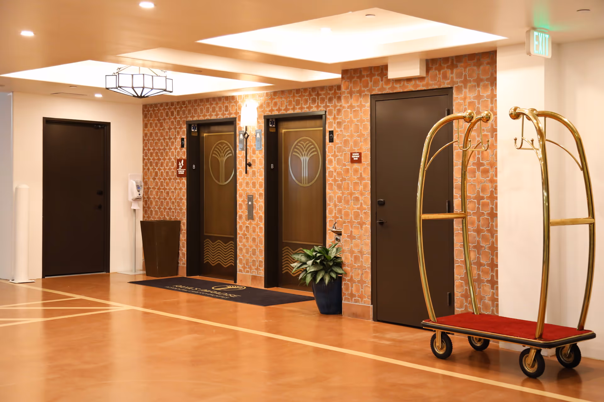 Interior hallway of Raya's Paradise San Clemente featuring two elevators with decorative doors, a luggage cart with a red carpet base, a potted plant, and brown doors against a patterned orange wall. Ceiling lights and an exit sign are also visible.