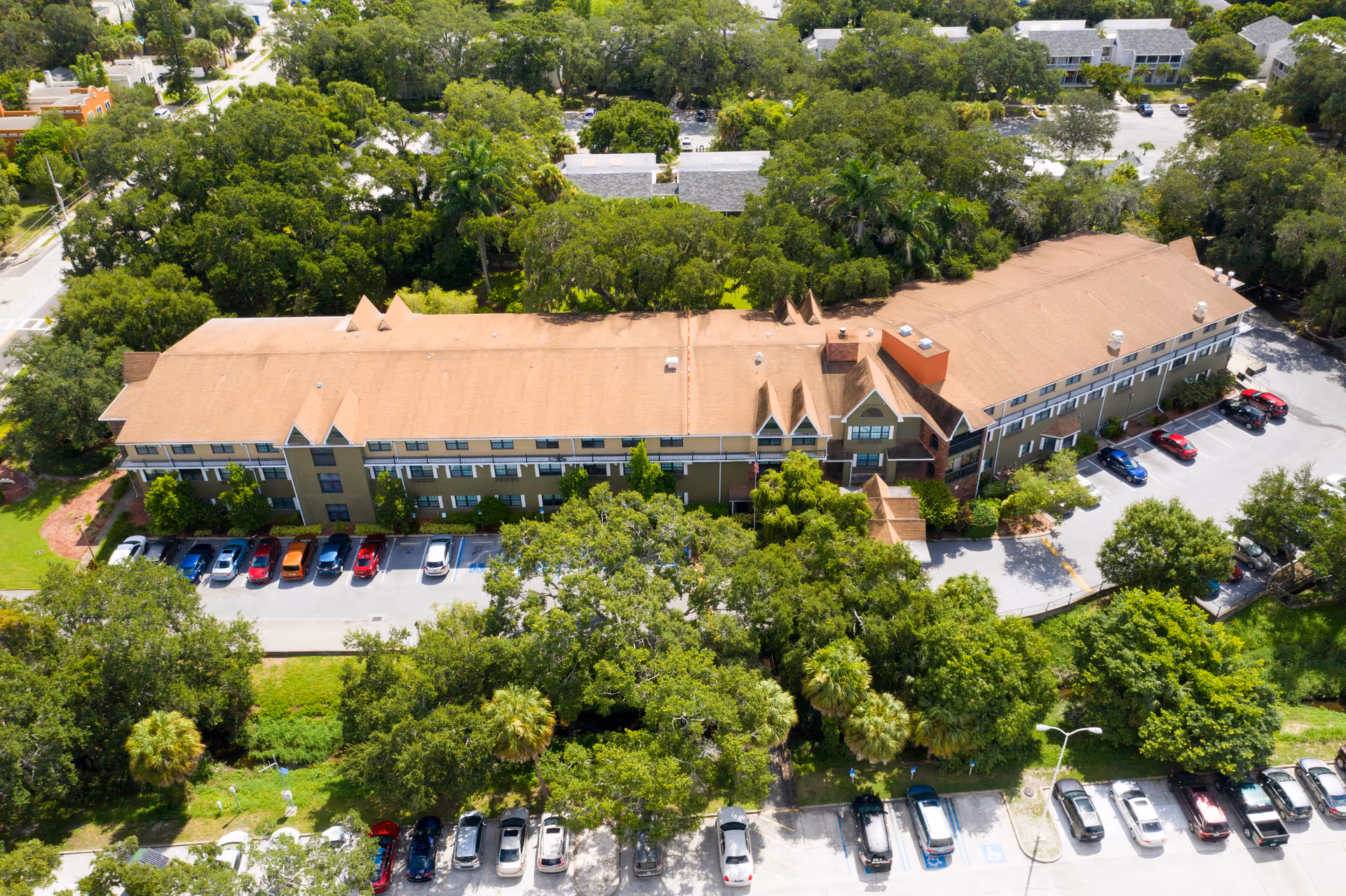 Aerial view of a long two-story building surrounded by trees with cars parked in front and rear parking areas.