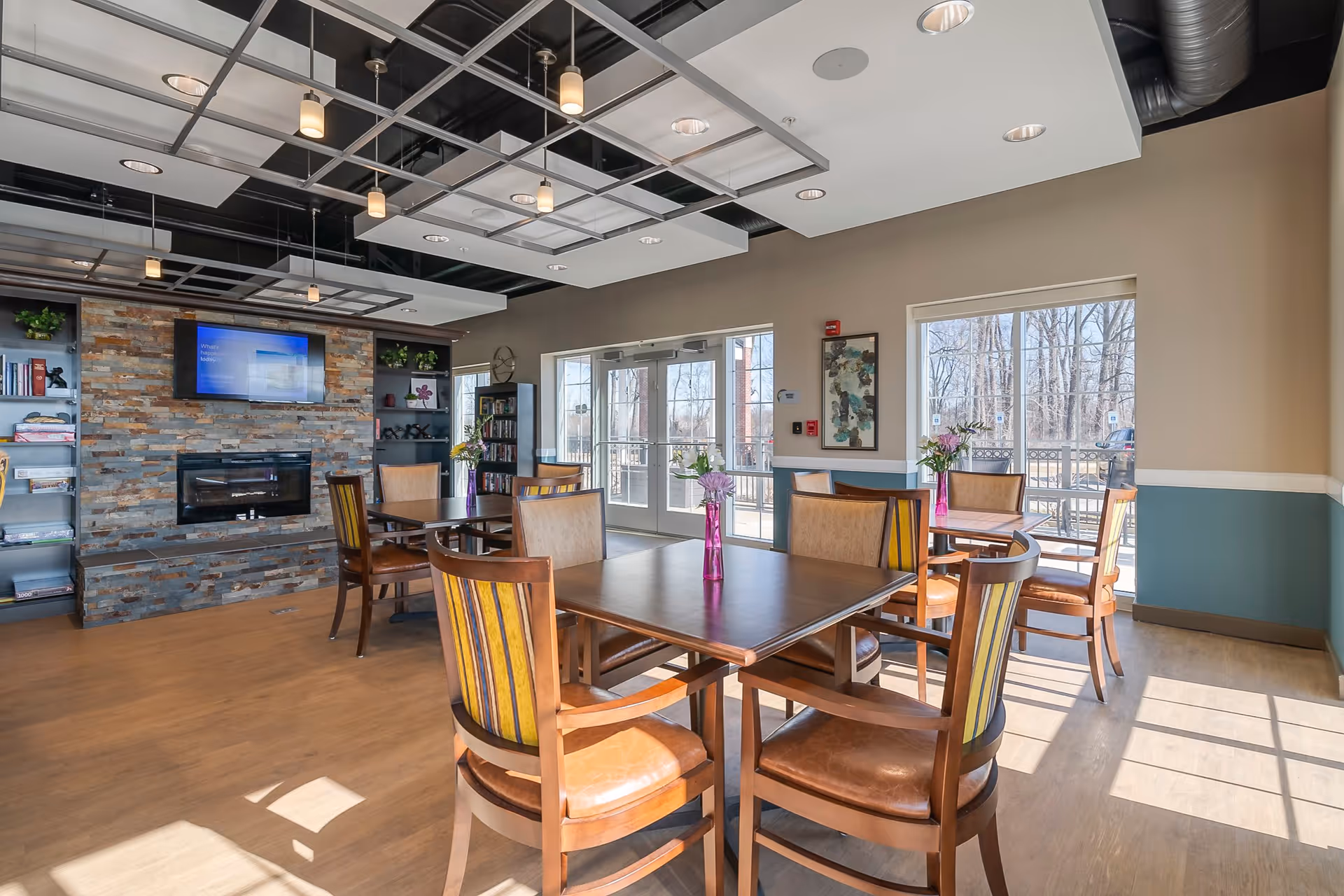 A bright and spacious common area with wooden tables and chairs arranged for seating. The room features large windows letting in natural light, a stone accent wall with a mounted flat-screen TV above a modern fireplace, and built-in shelves with books and decorative items. The ceiling has a modern design with hanging pendant lights.