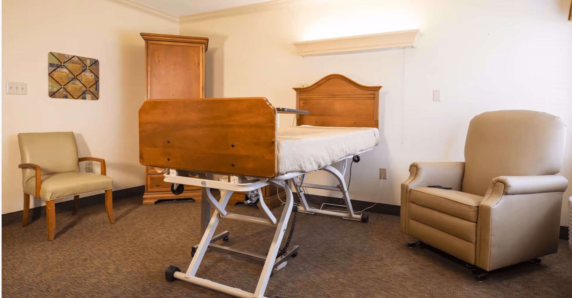 A room with a medical bed featuring wooden head and footboards, a beige recliner chair, a green armchair with wooden arms, a wooden armoire, and a decorative wall hanging. The room has beige walls and carpeted floor.