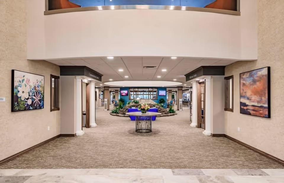 Spacious and well-lit interior hallway of a retirement community with beige walls, carpeted floor, and decorative paintings on both sides. A round table with a flower arrangement is centered in the hallway, with a stone water feature and greenery visible further inside. The ceiling has recessed lighting and a circular design element.
