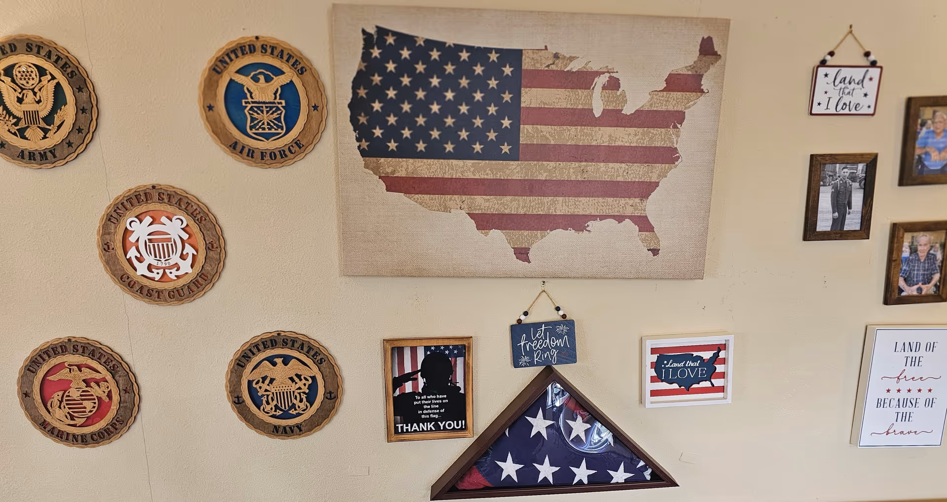 A wall decorated with patriotic and military-themed items including wooden plaques of United States military branches, a map of the USA with the American flag pattern, framed photos, a folded American flag in a display case, and signs with patriotic messages such as 'Let freedom ring' and 'Land of the free because of the brave.'
