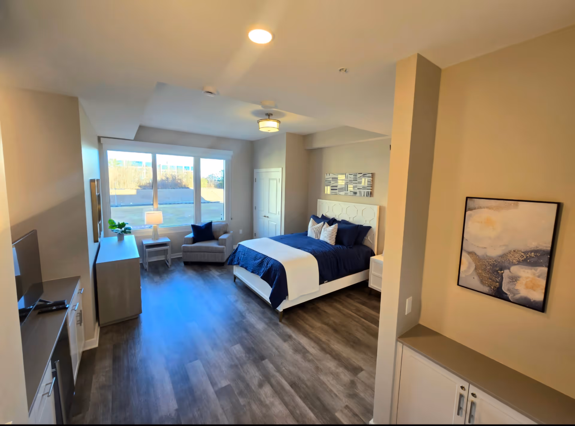 A bright and spacious bedroom with a large window letting in natural light. The room features a bed with navy blue bedding and white pillows, a comfortable armchair by the window, a dresser with a lamp and plant, a flat-screen TV on a cabinet, and a piece of abstract artwork on the wall. The flooring is wood-style and the walls are painted beige.