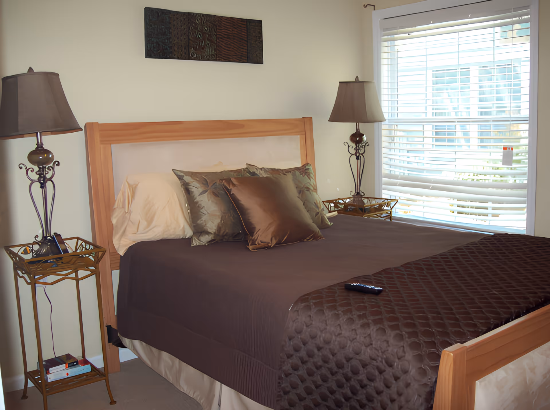 Neatly made bedroom with a wooden bed, brown bedding and pillows, matching bedside tables and lamps, and a window with blinds.