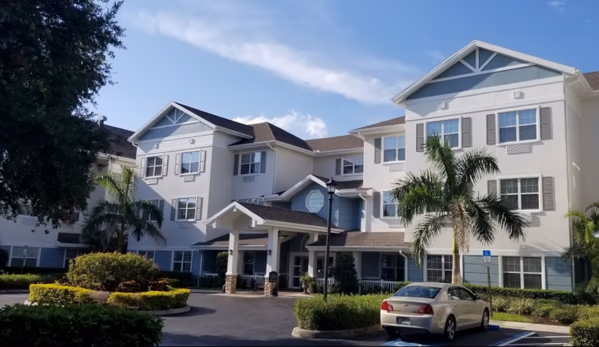 Exterior view of a three-story senior living facility building with white and blue walls, multiple windows, palm trees, and a car parked in a handicapped parking spot in front of the entrance under a covered driveway.