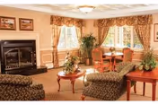 A cozy living room area with two patterned armchairs and a matching sofa arranged around a wooden coffee table with a floral centerpiece. There is a fireplace on the left side with framed pictures above it. Two large windows with floral curtains allow natural light to fill the room. A small round dining table with four chairs is positioned near the windows, and there are several potted plants adding greenery to the space.