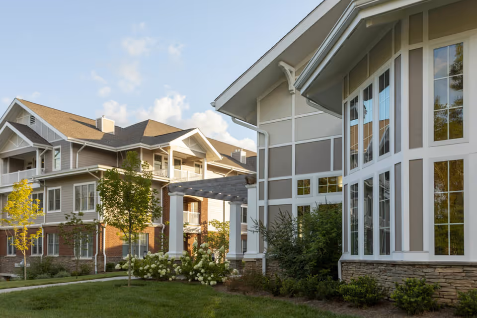 Exterior view of a senior living facility building with large windows, beige and white siding, stone accents, and a well-maintained lawn with trees and shrubs under a partly cloudy sky.