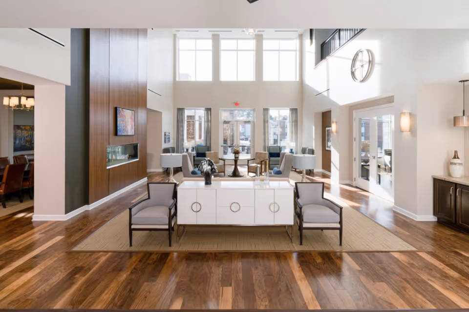 Bright and spacious living room area in a retirement community with high ceilings and large windows letting in natural light. The room features wooden flooring, a modern white console table with decorative items, two gray armchairs, a seating area with multiple chairs and tables, a wall-mounted TV above a built-in fireplace, and a large clock on the wall near glass doors leading outside.