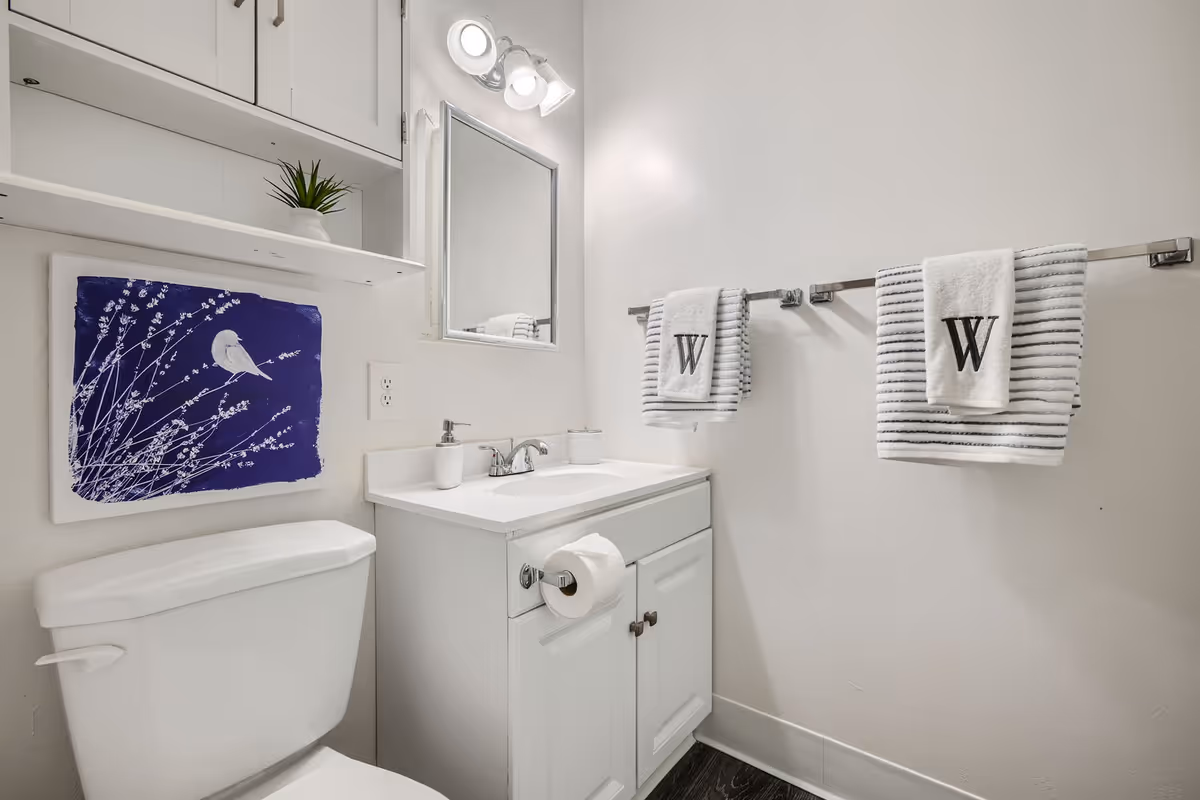 Small white bathroom with a toilet, vanity and mirror, wall shelves, blue bird artwork, and striped towels monogrammed with a 'W'.