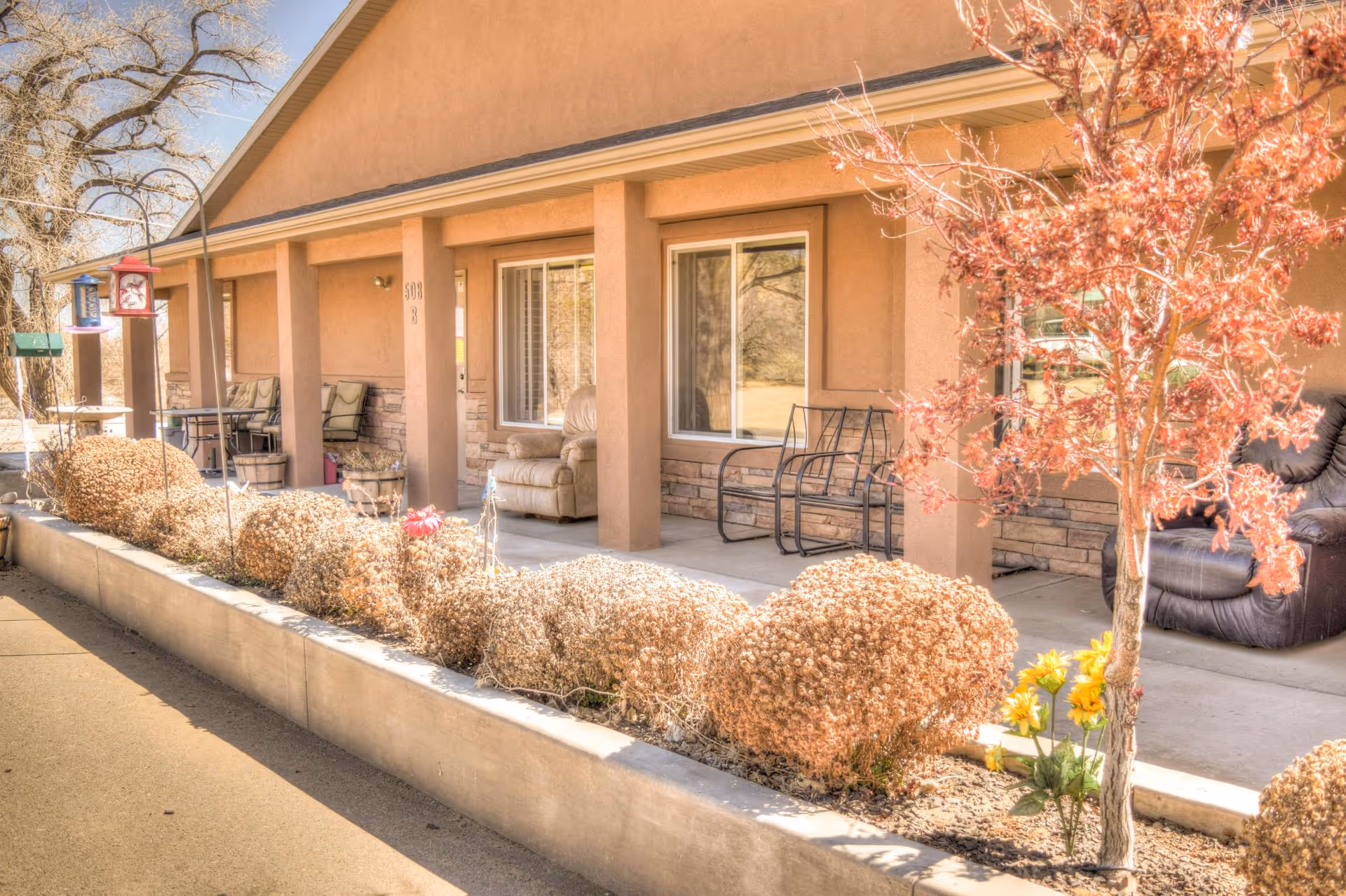 Outdoor view of a senior living facility porch area with cushioned chairs and small tables. There are trimmed bushes and a small tree with red leaves in front of the porch. The building has a beige exterior with stone accents and large windows.