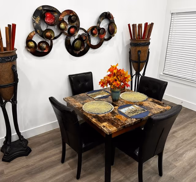 A small dining area with a square marble-top table set for four with decorative plates and placemats. Four black cushioned chairs surround the table. A vase with orange and red autumn leaves is centered on the table. On the wall behind the table is a modern metal wall art piece with circular designs in black, red, and gold tones. Two tall decorative urns with wooden sticks stand on either side of the table. The room has wood flooring and a window with white blinds.
