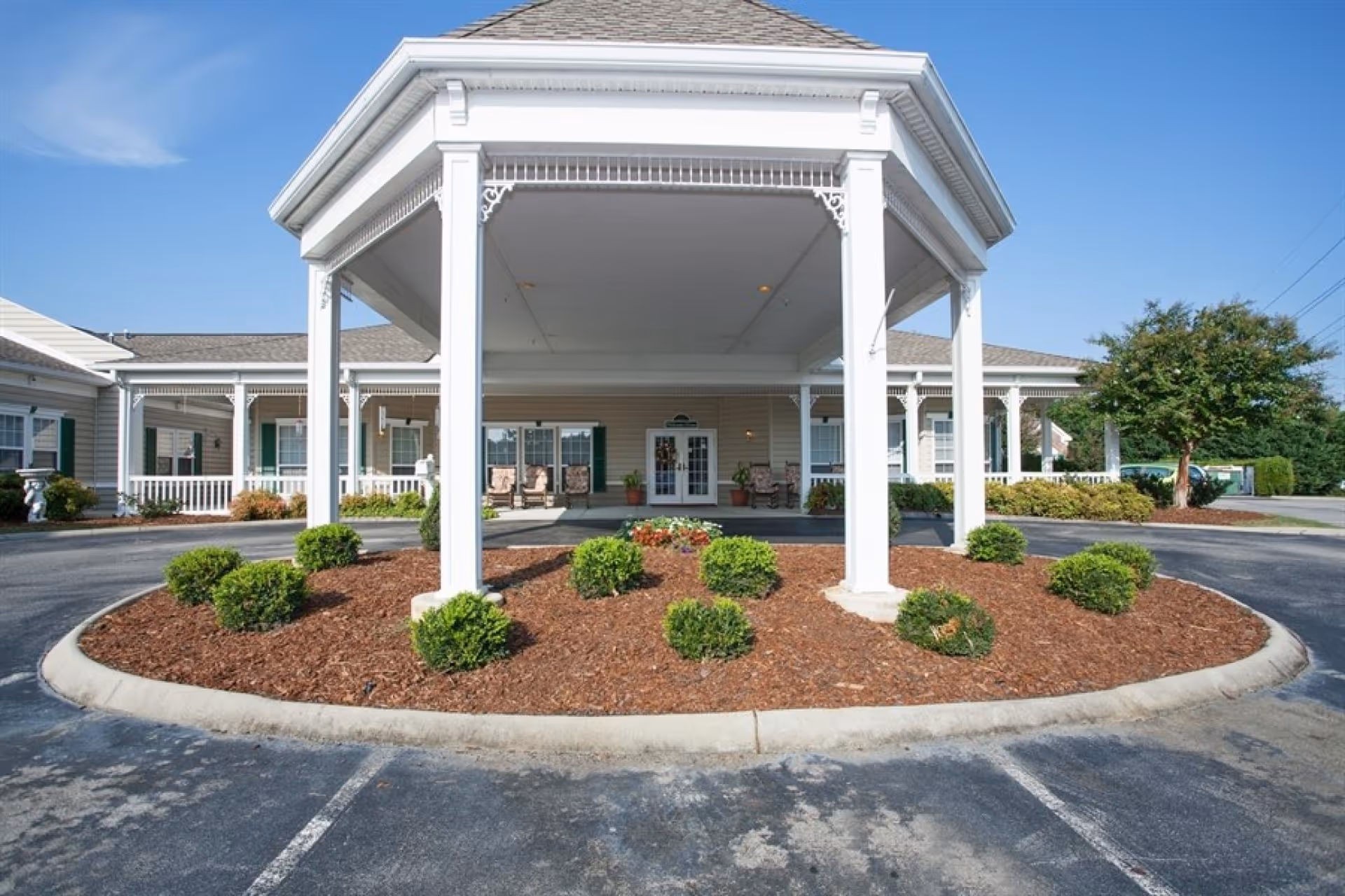 Covered circular driveway and main entrance of a single-story senior living facility with white columns, seating, and landscaped shrubs.