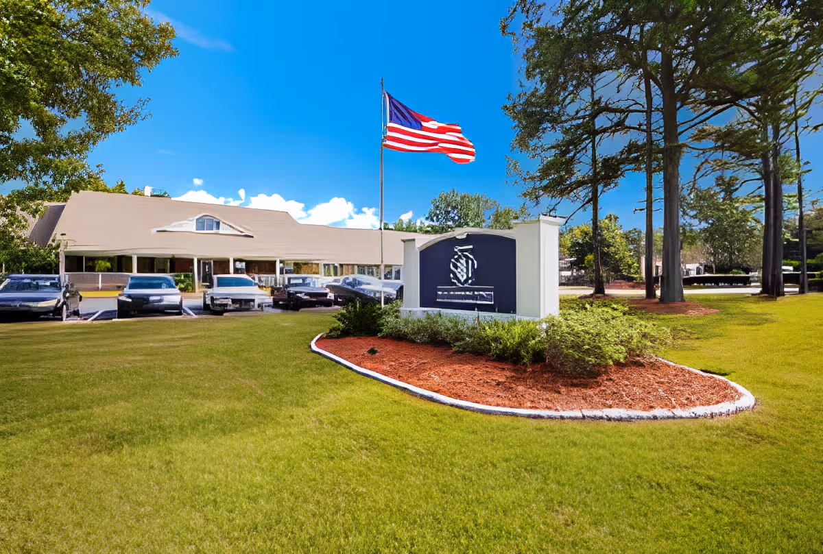Front exterior of a senior living facility showing a landscaped lawn, entrance sign, an American flag on a flagpole, and parked cars.