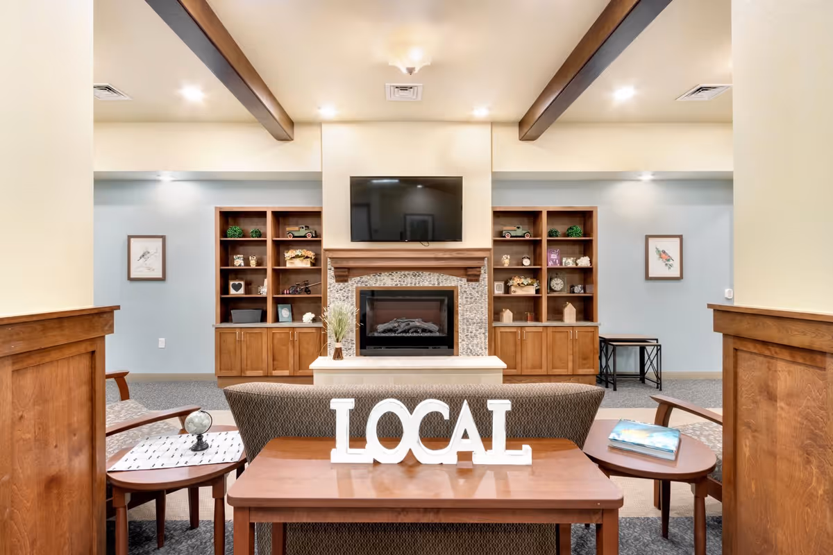 A cozy living room area with a fireplace and a mounted flat-screen TV above it. On either side of the fireplace are wooden shelves with decorative items and plants. In front of the fireplace is a sofa with a wooden table behind it displaying the word 'LOCAL' in white letters. There are side tables with a globe and a book, and the room features wooden beams on the ceiling and soft lighting.