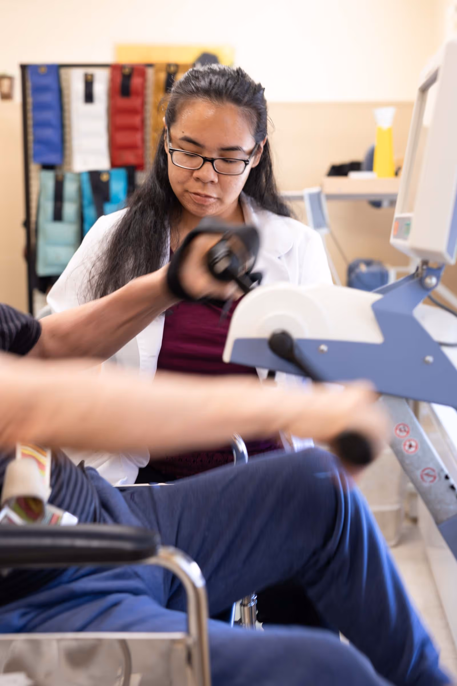 A healthcare professional wearing glasses and a white coat assists a person in a wheelchair who is using an exercise machine in a clinical or rehabilitation setting.
