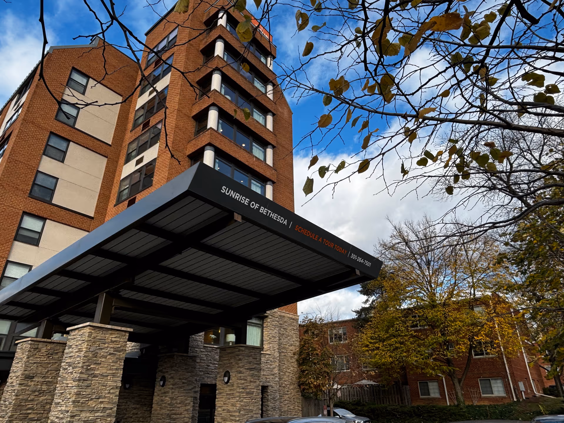 Exterior view of a multi-story brick building with a covered entrance canopy displaying the text 'Sunrise of Bethesda | Schedule a Tour Today | 301-264-7501'. Trees with autumn leaves and a partly cloudy sky are visible around the building.
