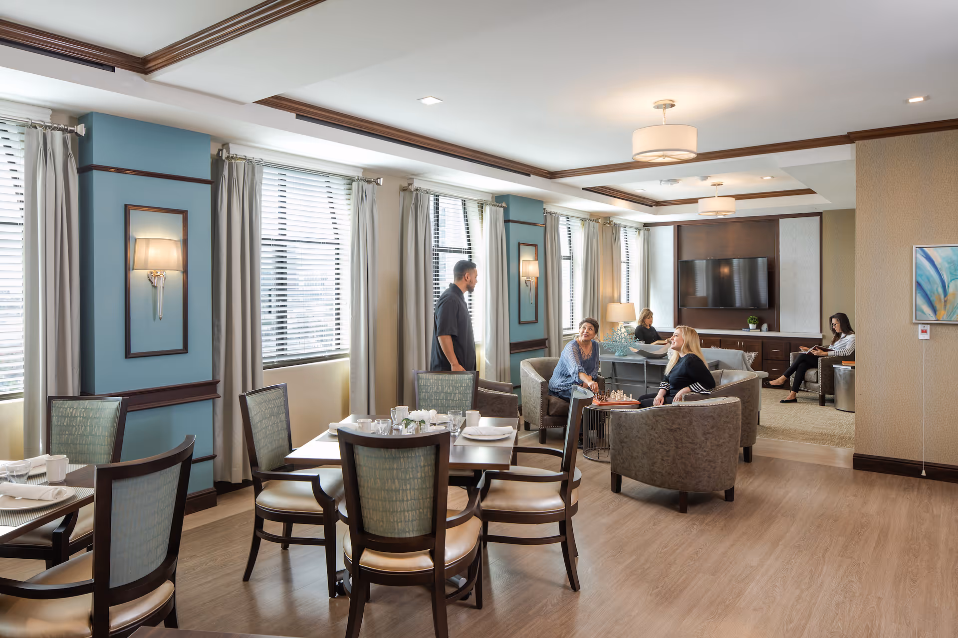 A bright and spacious common area in a senior living facility with several people sitting and conversing. The room features large windows with white blinds and beige curtains, light blue walls with wooden trim, and modern ceiling lights. There are dining tables and chairs in the foreground and a seating area with armchairs and a television mounted on the wall in the background.
